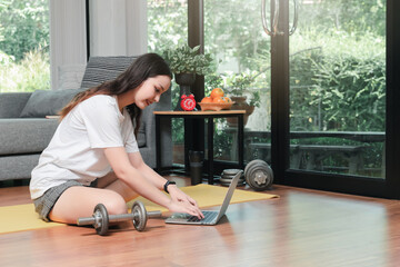 Beautiful young woman with dumbbell watching fitness video tutorials online on laptop while sit...