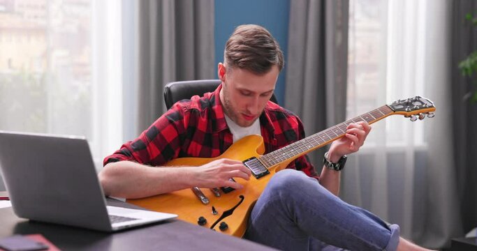 A male student is learning to play the electric guitar. A young boy reviews a webinar on guitar playing on a laptop while sitting in a chair at his desk in his room.