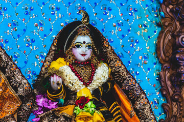 Close up of white marble stone statue of goddess Radha with beautiful clothing and jewellery, decorated with bright flowers in blue background at an Indian temple.