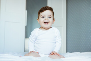Fototapeta premium A portrait of a cute toddler baby boy in a white bodyuit seating on bed and laughing.Happy baby face look at the camera, in bedroom at home