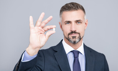 mature business man with grizzled hair in suit hold pill in hand with ok gesture, energy remedy