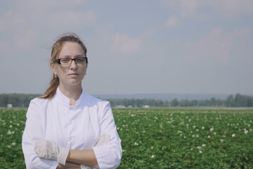 Middle shot portrait of attractive Caucasian woman in eyeglasses and white lab coat posing in field. Portrait of female agronomist or scientist standing in field, looking at camera. 