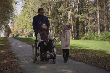 Happy family of three is walking in the autumn park, a father is driving a disabled mother in a wheelchair, little daughter walking nearby