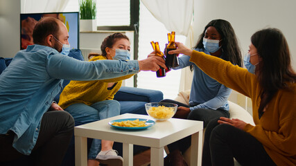 Fototapeta premium Happy diverse young people having fun in living room, clinking bottles of beer telling stories and jokes during global pandemic. Multiethnic group of friends celebrating with toast in outbreak