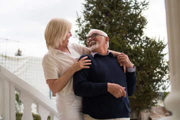 A good-looking senior couple is standing on a porch, talking and smiling. The wife stands on the...