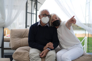 A good-looking senior couple is sitting on a balcony with the protective face masks on. The wife is leaning on her husband's shoulder.