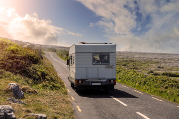 White camper van on a small asphalt road leading into sun. Traveling in motor home concept. Warm sunny day. Burren area, Ireland. Sun flare. Summer vacation concept