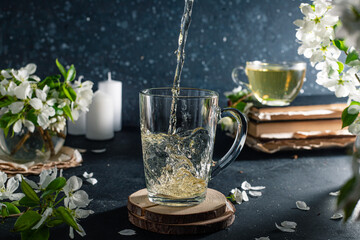 flower tea in a transparent mug on a dark background. Next to the cup are books and branches with white apple blossoms. The tea spills out of the mug