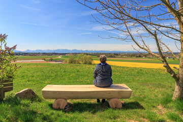 Frau im mittleren Alter sitzt alleine auf einer Holzbank mit Blick auf die Alpen