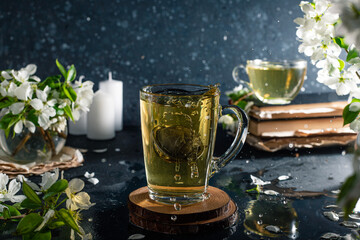 flower tea in a transparent mug on a dark background. Next to the cup are books and branches with white apple blossoms. The tea spills out of the mug