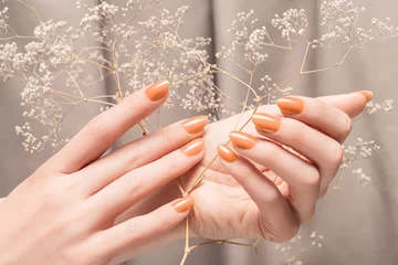 Poster Female hands with glitter beige nail design. Female hands hold autumn flower. Woman hands on beige fabric background © devmarya