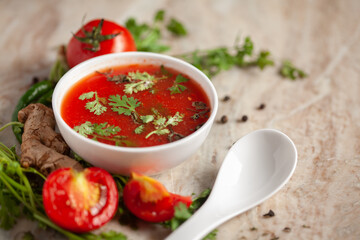 Close-up of Indian Homemade fresh and healthy tomato soup garnished with fresh coriander leaves and ingredients and herbs,  served in a white ceramic bowl with a spoon over marble top. 