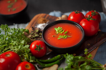 Close-up of Indian Homemade fresh and healthy tomato soup garnished with fresh coriander leaves and ingredients and herbs, served in a black bowl over the wooden top background. 