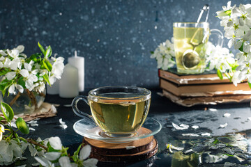 flower tea in a transparent mug on a dark background. Next to the cup are books and branches with white apple blossoms
