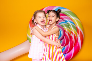 two happy little girls in colorful dress laughing hugging having fun on yellow background with lollipop
