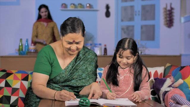 Caring Indian grandmother helping her grandchild in completing homework. Young mother busy working in the kitchen while an old woman taking care of her girl - family care and relationship