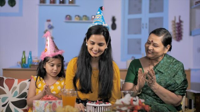 A young woman with a party cap cutting her birthday cake sitting with family. Medium shot of cheerful Indian lady celebrating women's day with her daughter and mother - family bonding and care