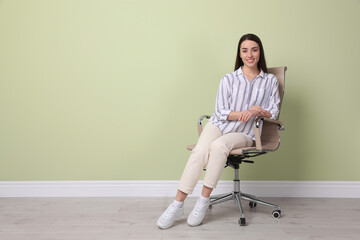 Young woman sitting in office chair near green wall indoors, space for text