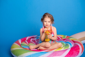 cute child girl in swimming suit drinking orange juice sitting on colorful inflatable mattress on blue background.