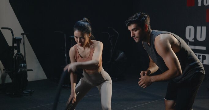 Asian young beautiful woman doing exercise sweat extreme with battle rope in cross fit gym training at club fitness GYM with trainer motivating man, health workout sport trends concept