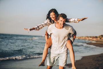Happy smiling summer couple piggyback together with arms outstretched at beautiful beach
