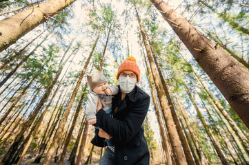 Naklejka na ściany i meble portrait of attractive nerd in long sleeve black coat and orange hat with glasses and protective face mask with baby in arms outside in the pine forest at sunset