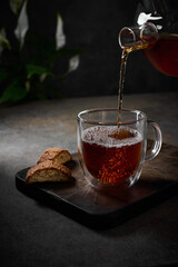 Black tea is poured from the teapot into transparent cup with bubbles. Nearby cookies on cutting board. Selective focus. Vertical orientation, low key, rustic