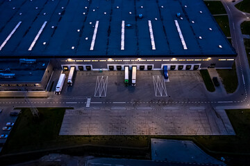 Aerial view of goods warehouse at Night. Logistics center in industrial city zone from above. Aerial view of trucks loading at logistic center. View from drone.