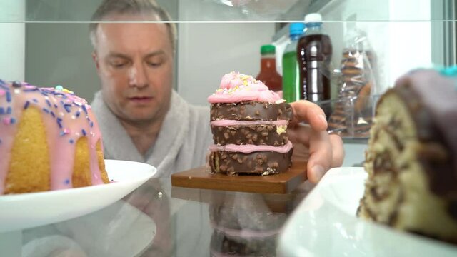 Man Opening Fridge Door And Taking Out A Piece Of Cake And A Lot Of Sweets.