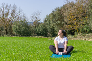 Curvy woman practicing yoga on a mat in the grass looking up at the sun