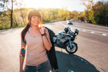 World Motorcyclist Day. Portrait of happy young tattoed woman with a leather jacket poses against the background of a motorcycle by the road. Motorcycle travel concept
