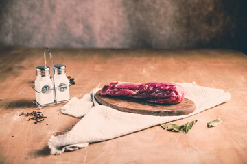 A piece of raw pork tenderloin on a chopping board with seasonings on the table