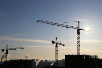 Silhouettes of construction cranes on blue sky and sunshine background. Housing construction in city