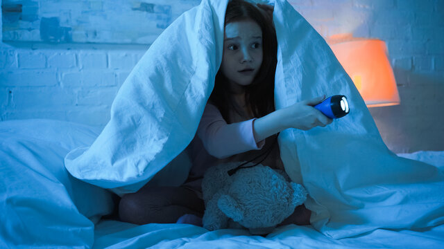 Scared Kid With Teddy Bear Holding Flashlight On Bed In Night