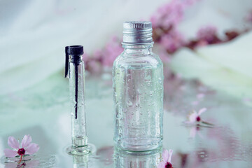 Closeup of wet glass flasks and blurred tender pink flowers on the background with wet reflecting surface underneath. Herbal medicine and cosmetics concept.