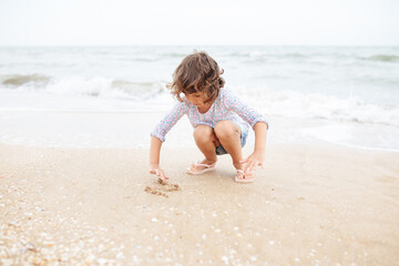 Fototapeta na wymiar Five years cute curly caucasian girl painting on sand on the beach.