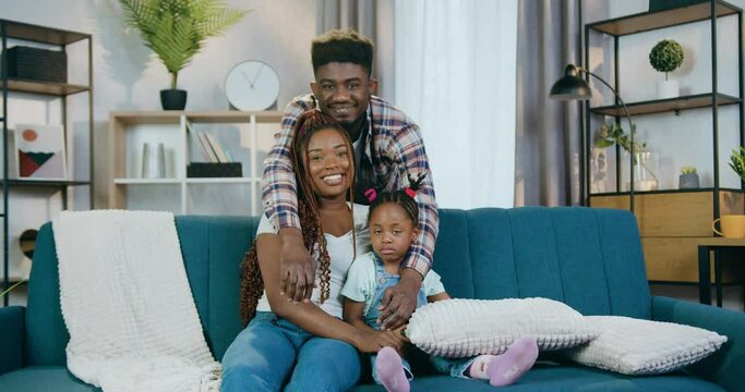 Beautiful black woman sitting on couch with pretty baby while handsome man standing behind and embracing them. Mother and father enjoying time spending with their daughter.