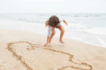 Five years cute curly caucasian girl painting on sand on the beach.