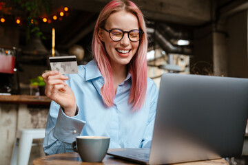 Young laughing woman using laptop and credit card while sitting in cafe