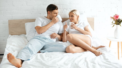 smiling young woman looking at boyfriend drinking coffee in bed