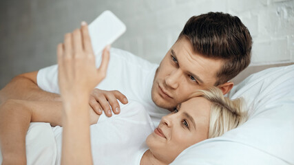 young man and joyful woman taking selfie on smartphone in bedroom