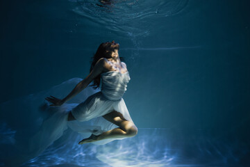 Fototapeta na wymiar lighting on young graceful woman in white elegant dress swimming in pool with blue water