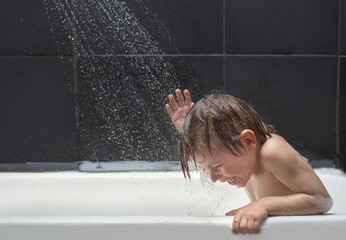 the child is sitting in the bath, water is pouring on him from the shower, splashes are flying, he...