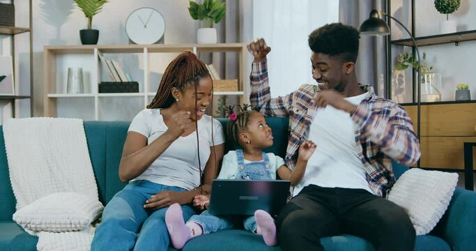African parents and cute daughter using wireless laptop for playing online games while staying at home. Happy family sitting on couch and gesturing for express their feelings of victory.