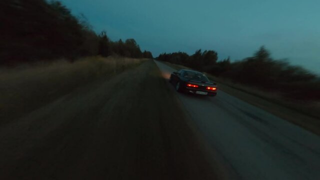 Sport black jdm vehicle drives along empty narrow rural road and does police turn drifting at forest in dark evening first point view cinematic aerial shot by fpv racing drone.