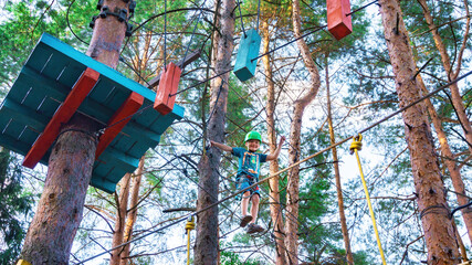 A boy in a helmet boldly overcomes a difficult section along a rope ladder stretched between trees in an adventure high-rope town. Ideas of sports activities for children during the summer holidays.