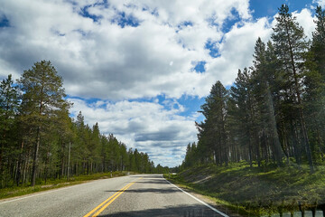 Fototapeta premium View from relief car windscreen on the blue sky with white clouds, grey asphalt road and landscape with forest and green teeses. Landscape through window
