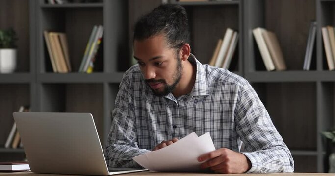 African businessman analyst sit at desk hold sheets check documents work on laptop, read contract papers contract, doing online research, pay bills use e-bank app, make resume, prepare report concept