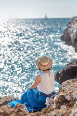 A girl in a hat sits on the seashore and looks into the distance. The woman is looking at the sea. Travel to the sea. High quality photo