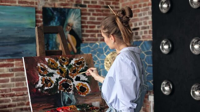 Young beautiful female artist painting still life with sunflower on canvas using oil paintings and art brush. Woman student at fine art course. Relaxation, leisure, hobby, stress management.